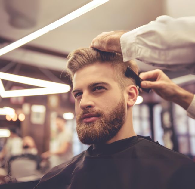 Homem loiro com barba, sentado na cadeira do barbeiro enquanto o barbeiro corta o seu cabelo