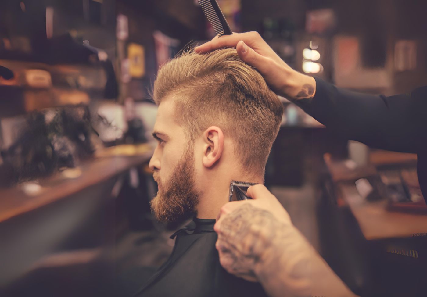 Homem loiro sentado na cadeira do barbeiro que faz acabamentos em sua nuca com máquina de cortar cabelo.