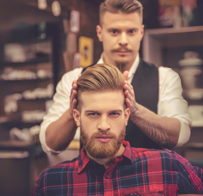 Homem loiro com barba e cabelo cortados, sentado na cadeira do barbeiro após serviço finalizado.